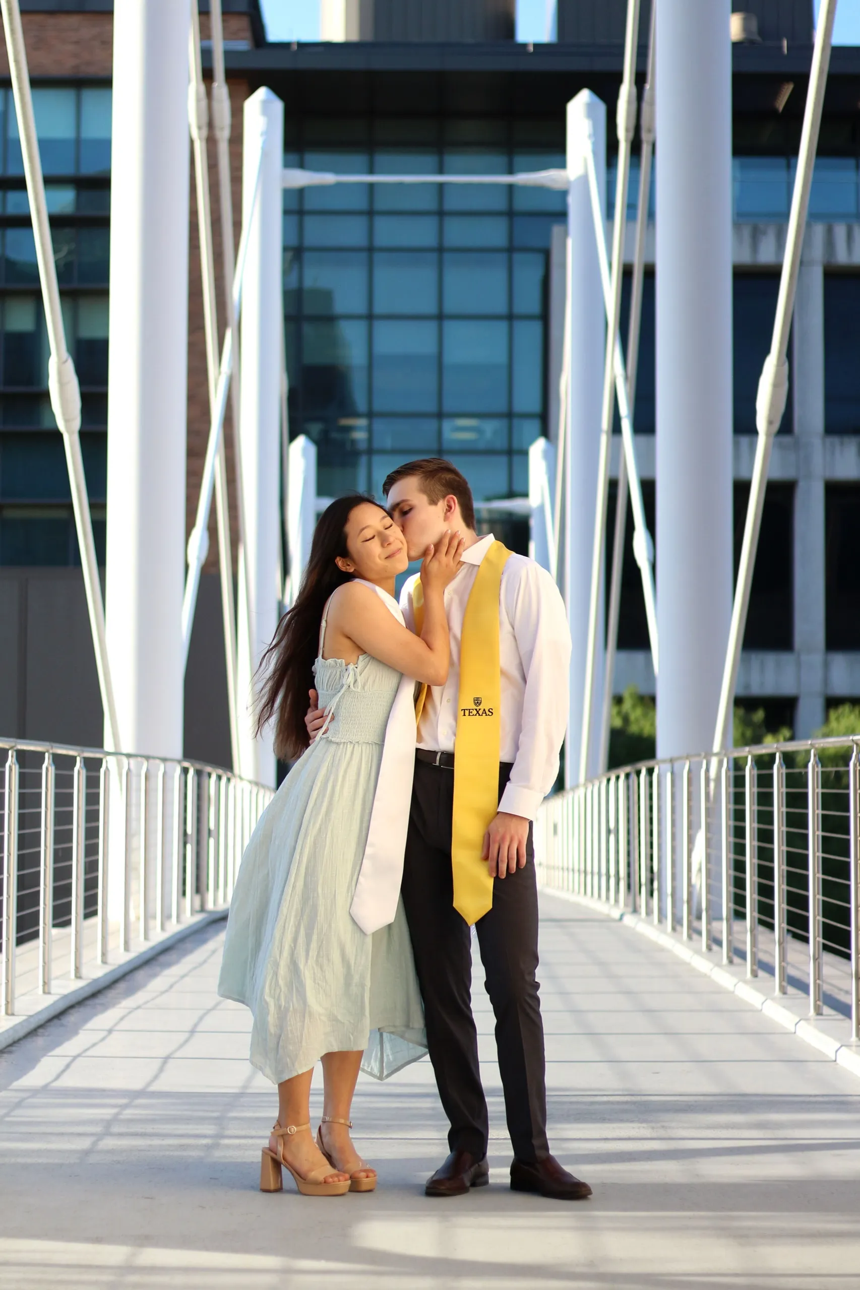 A graduate couple kissing on a white suspension footbridge, one of them wearing a gold Texas stole.