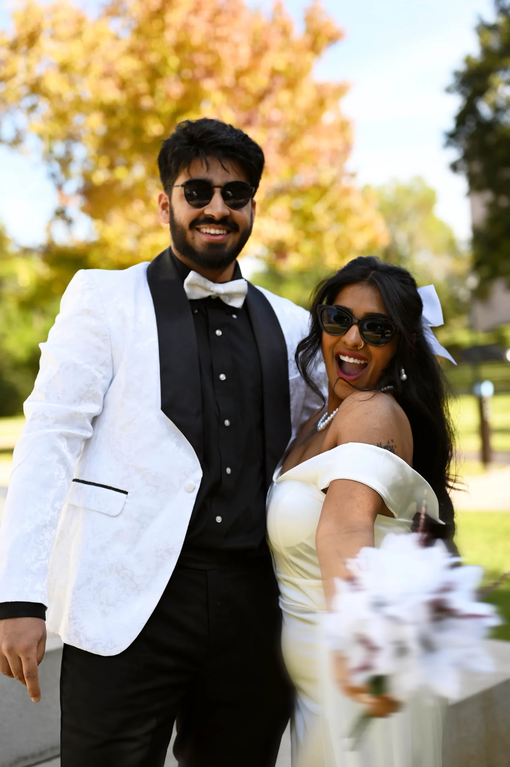 A groom in a white tuxedo jacket and a laughing bride in sunglasses holding a white bouquet, in front of autumn trees.