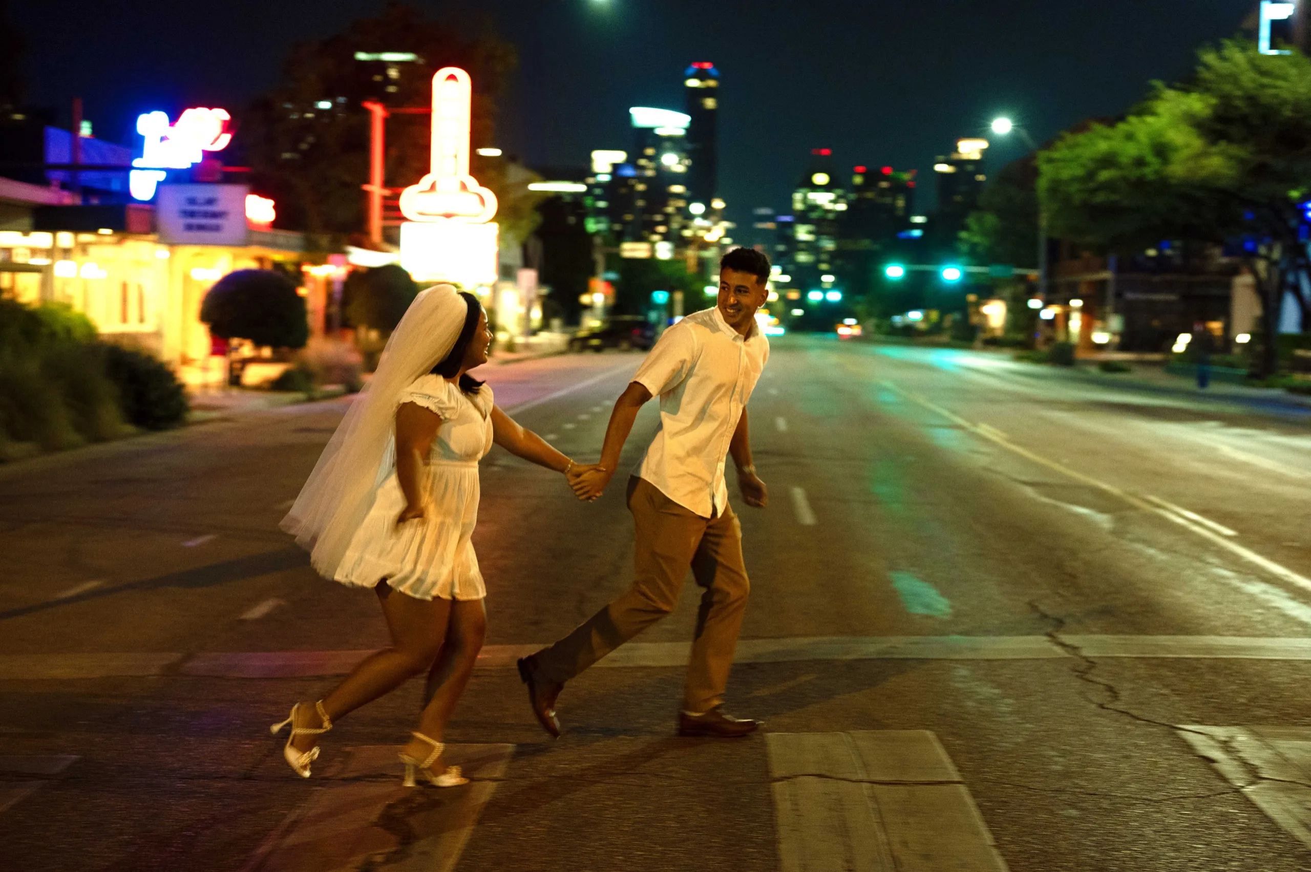 A bride in a short white dress and veil leads her partner by the hand across a downtown street at night, the skyline lit up behind them.