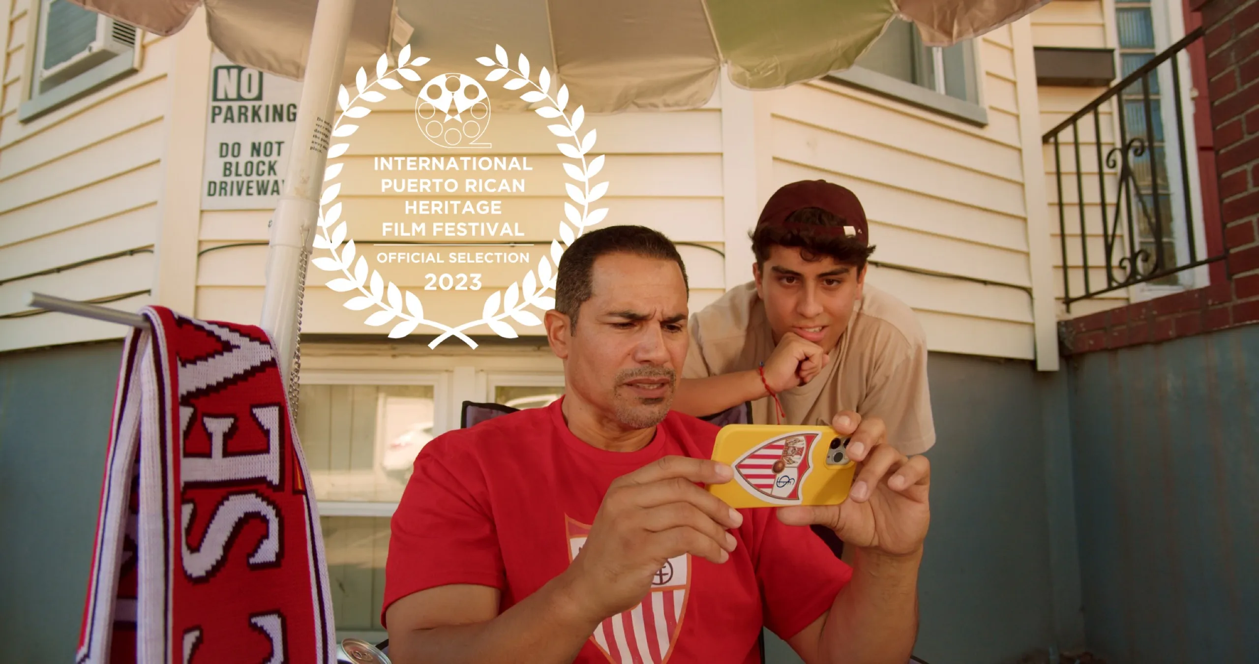 Film still from B-Line. A man in a red jersey looks at a phone on a porch while a younger man leans over his shoulder.